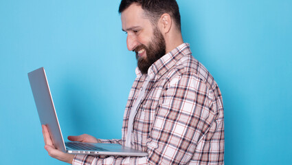 Studio portrait of young bearded man standing holding laptop and looking at camera with happy smile isolated on blue background. 