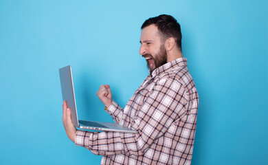 Happy excited Caucasian bearded man holding laptop and raising his arm up to celebrate success or achievement on blue background. 