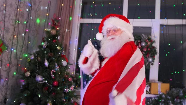 Close-up of gray-bearded Santa wearing American flag and dancing against backdrop of Christmas Tree decorated with toys and sparkling garlands. New Year party.