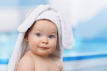 Portrait happy baby girl in white towel with ears after swimming lessons in pool, blur background