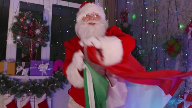 Positive man in Santa Claus costume with Italian flag, with gray beard and glasses, dances in festively decorated house next to Christmas tree and twinkling garlands.