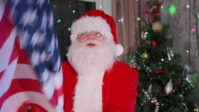 Santa Claus waving American flag, symbol United States, close up, at festive Xmas party, backdrop of Christmas tree and tinsel. USA support. 
