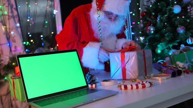 Laptop with chromakey screen on table against background of Santa Claus wrapping gifts, present boxes, Christmas tree. During New Year. Close up. Xmas preparing.