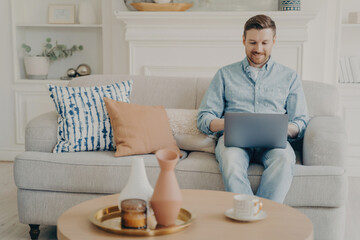 Man in denim shirt using laptop on sofa, comfortable home living with coffee on table