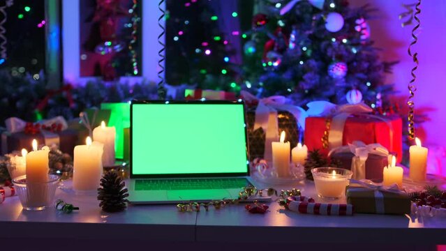 Close up, laptop green screen place promo ads offer, night time, among flickering garlands, xmas tree, flaming candles, cones, and tinsel. Marry Christmas and Happy New Year holidays background.