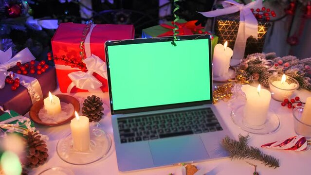 Static laptop with green chromatic screen close-up, xmas decor, burning candle, gift boxes, tinsel and fir tree. Advertising platform. Christmas background. 