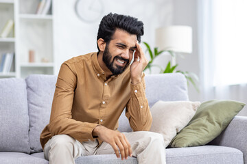 Young Indian man suffering from headache and pressure. Hiding, he sits on the sofa at home, holding his head with his hand, overtired.
