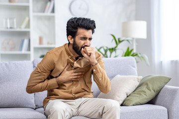 Indian young man sitting at home on the couch and coughing, covering his mouth, holding his chest,...