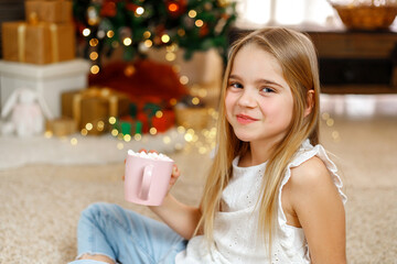 Little girl with a pink mug of cocoa, near the Christmas tree on Christmas Eve at home.