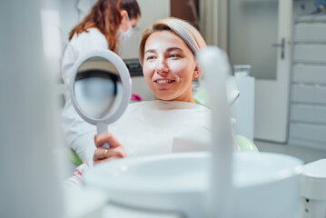 Portrait of young smiling woman sitting in stomatology clinic chair and looked at mirror evaluating...