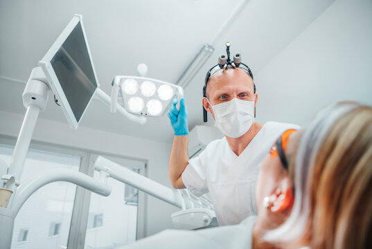 Dental clinic patient appointment in modern medical ward. Dentist doctor in magnifying glasses pointing light at young Female ready for teeth surgery. Health care and medicare industry concept image