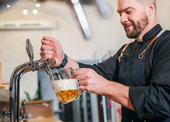 Smiling stylish bearded barman dressed black uniform with an apron tapping fresh lager beer into glass mug at bar counter. Successful people, beer consumption, beverages industry concept image