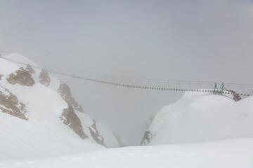 Suspension bridge between two mountain peaks in gloomy weather with fog and snow