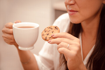 woman tastes crisp cookie with chocolate chip and drink hot tea at home