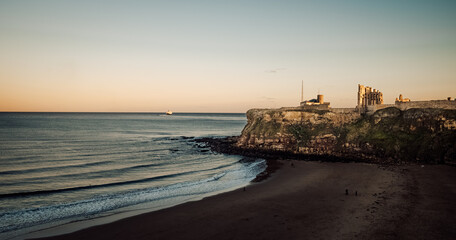 As the sun rises over the wild ocean, a lone boat floats towards the distant horizon, framed by the rugged cliff and sandy shore of the untamed beach