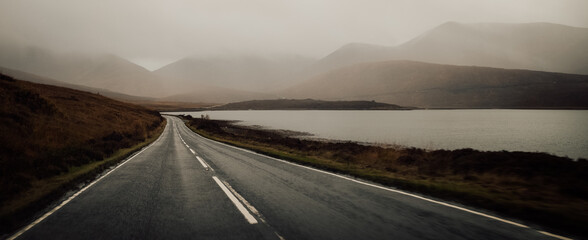 A winding highway cuts through the misty highlands, leading to a tranquil lake nestled between towering mountains and a vast open sky, inviting travelers to embark on a journey of discovery and awe i - obrazy, fototapety, plakaty