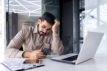Upset sad man at workplace inside office, businessman uses phone reads bad news received by e-mail, employee disappointed holds smartphone in hands browses internet sites and social networks. - Powered by Adobe