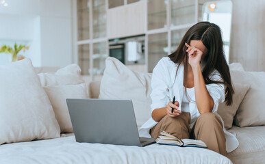 tired brunette is sitting on the couch at home working with a laptop and with her diary open, she covers her face and touches her forehead, exhausted, Crying lady, grief. - obrazy, fototapety, plakaty
