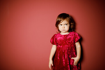 Pretty caucasian brunette toddler little girl in red velour dress dissatisfied and angry at the red background. Christmas vibe 