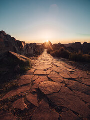A stunning madeira sunrise reveals a golden stone path, leading through a rugged landscape of mountains and desert, illuminated by the warm glow of the sun and stretching towards the infinite horizon