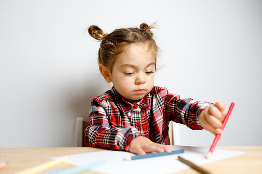 Portrait of cute toddler girl with two bunches in checkered dress sitting on the baby wooden table and chair and drawing with colorful pencils, Montessori concept fine motricity 