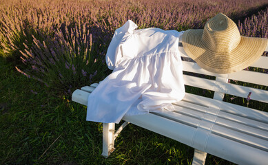 A woman's straw hat lies on a white wooden bench, a bouquet of lavender lies on it, the sunset in the rays. Place for writing, background	
