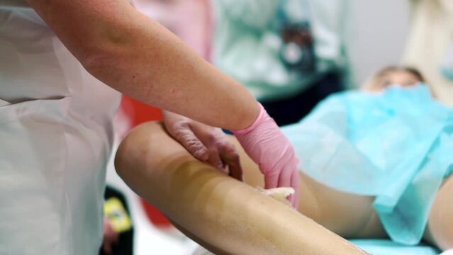 The hands of a master in a white coat and pink latex gloves manually remove sugaring paste from a bare female leg in a beauty salon. Hair removal and depilation for the beauty of the female body