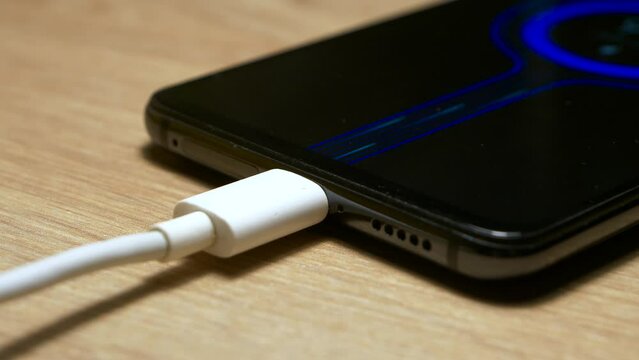 USB charging cable connected and transferring power to smartphone, with quick charge blue fluid particles animation on the screen. Black color modern smartphone, lying on a wooden table while charging
