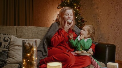 Mother with a toddler sitting on a couch and eating gingerbread cookies, having holiday time...