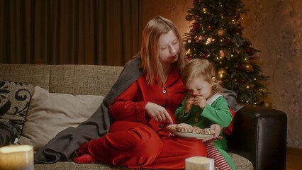 Mother with a toddler sitting on a couch and eating gingerbread cookies, having holiday time together. Woman in red pajamas and Christmas tree in the background plays with a child.