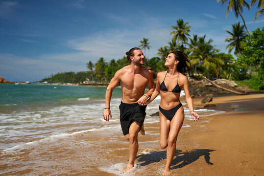 Active couple runs on tropical beach. Fit man and slender woman in swimwear enjoy seaside jog. Athletic duo sprints by ocean, embodying healthy lifestyle. Vacation exercise, sunny travel escape.
