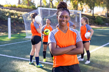 Young African American woman standing with a soccer ball in front of afFemale soccer team. The women players work together as a team in a group of five wearing matching orange jerseys. 