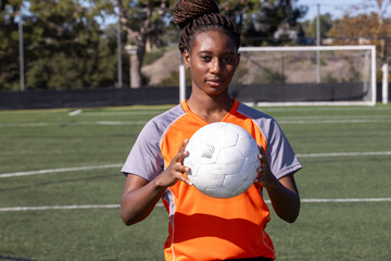 Young African American female soccer player holding a ball on the field. Her black hair is in braids and tied back. 