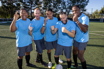 Soccer players wearing blue jerseys. The team is celebrating and the men are happy shouting and giving high fives after a score during a game. 