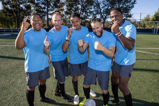 Soccer players wearing blue jerseys. The team is celebrating and the men are happy shouting and giving high fives after a score during a game. 