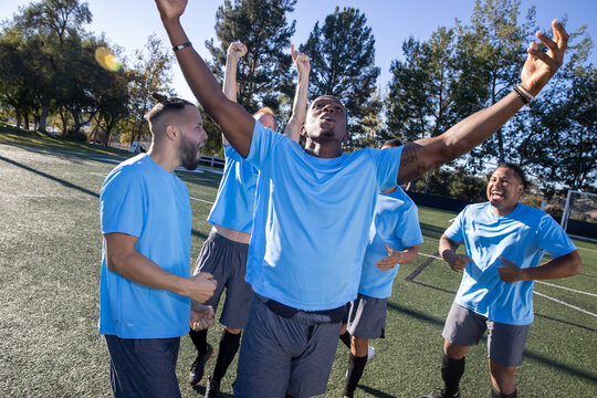 Soccer players wearing blue jerseys. The team is celebrating and the men are happy shouting and giving high fives after a score during a game. 