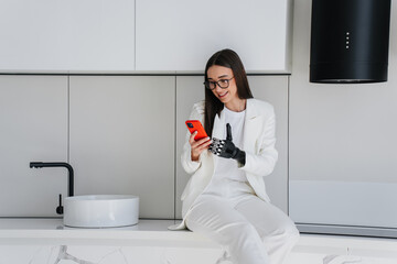 Cheerful European brunette girl holds phone using hi tech artificial arm sitting at kitchen...