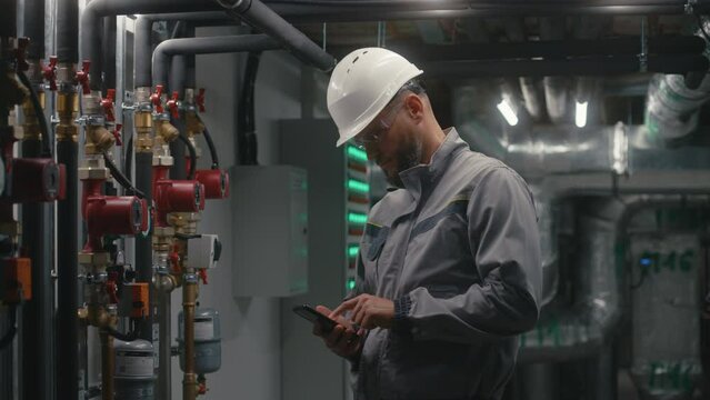 Male technician worker wearing safety uniform, protective glasses and hard hat works on modern plant, uses phone. Professional engineer maintains modern manufacturing factory or industrial facility.
