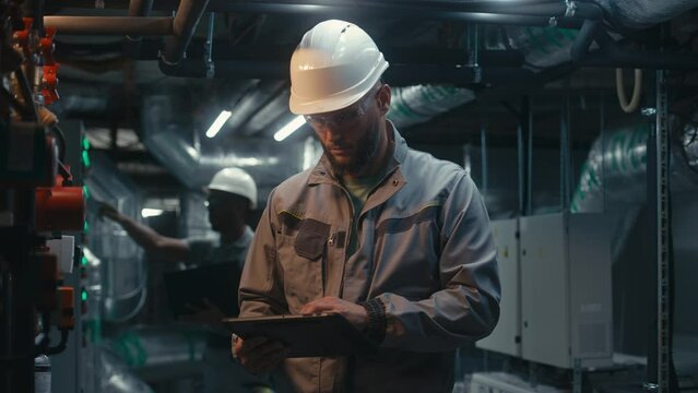 Diverse team of factory specialists work on modern plant using laptop and tablet computers. Industrial workers wearing safety uniform and hard hat inspect pipeline system. Heavy industry concept.