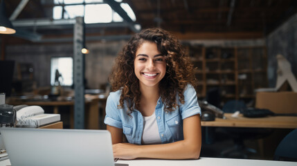 Cheerful woman sitting at a desk with a laptop in a warehouse surrounded by shelves stocked with boxes. - obrazy, fototapety, plakaty