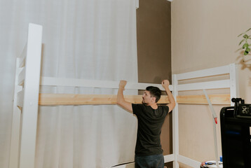 A young man connects a board into a hole on the two side of a bunk bed.