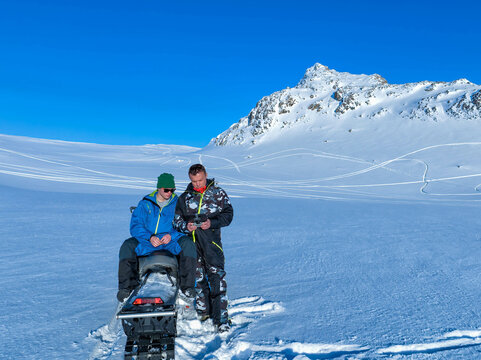 Son and father looking at drone remote control while taking pictures in winter snowy mountains. Perfect sunny day, snowmobile. Sweden. Scandinavian Mountains