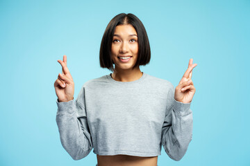 Smiling young asian woman wearing casual clothes with crossed fingers on blue background looking up