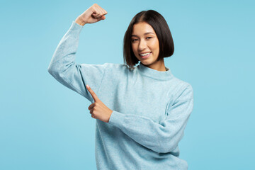 Young strong Asian woman wearing stylish sweater pointing finger at biceps looking at camera