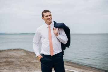 Handsome groom in a black suit with a red tie near sea. Wedding portrait of newlyweds.