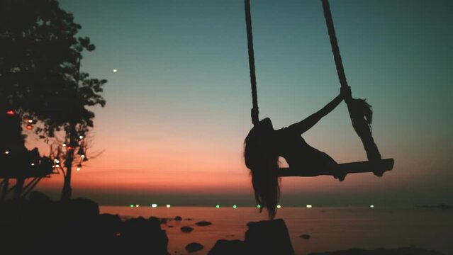 Happy girl swinging on swing in beach at sunset, bright colorful sky. Paradise sea relax, summer enjoyment, serene leisure, freedom, happiness. Outdoor lifestyle travel, holiday vacation. Slow motion