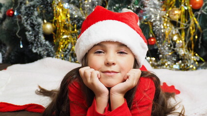 happy girl in santa claus hat hold hand demonstrate x-mas newyear advert promo wear warm winter clothes sings songs, congratulations a New Year's Christmas tree with toys and lights.
