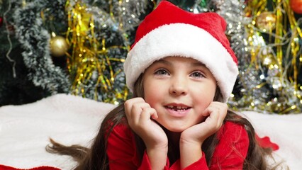 happy girl in santa claus hat hold hand demonstrate x-mas newyear advert promo wear warm winter clothes sings songs, congratulations a New Year's Christmas tree with toys and lights.