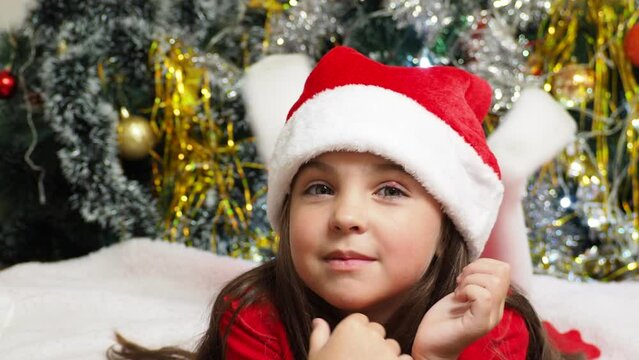 happy girl in santa claus hat hold hand demonstrate x-mas newyear advert promo wear warm winter clothes sings songs, congratulations a New Year's Christmas tree with toys and lights.