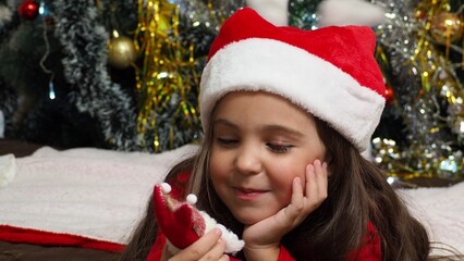 happy girl in santa claus hat hold hand demonstrate x-mas newyear advert promo wear warm winter clothes sings songs, congratulations a New Year's Christmas tree with toys and lights.
